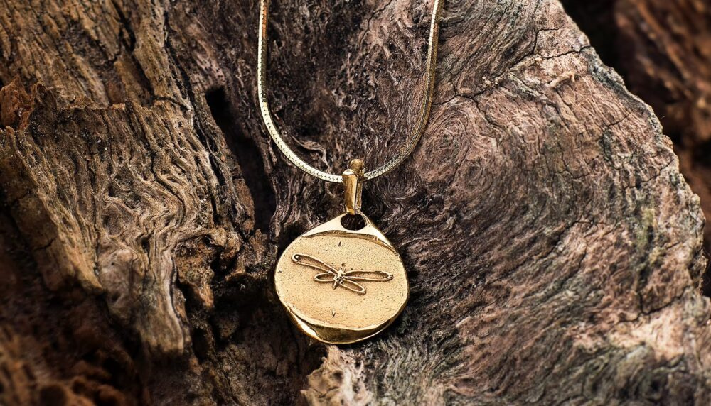 Gold necklace with a round pendant showing a dragonfly engraving, resting on rough brown wood.