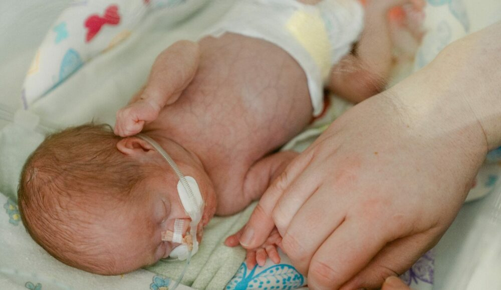 Mama ligoninėje švelniai laiko neišnešiotą naujagimį ant krūtinės.