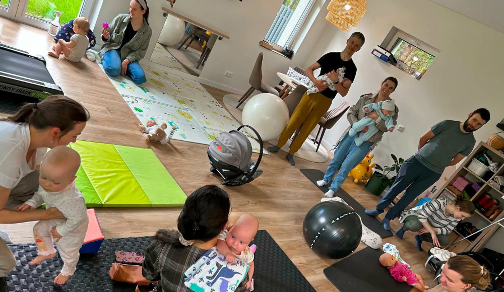 Group of caregivers and babies in a bright playroom with mats, toys, and a large exercise ball, all engaging with infants.