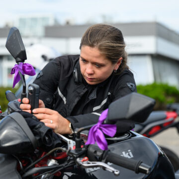 Woman in motorcycle gear leaning over a black bike, adjusting the handlebar; purple ribbons tied to the mirror.