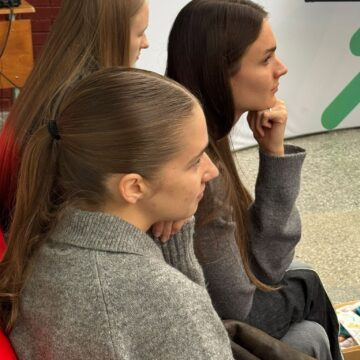 Three young women sit side by side indoors, wearing gray sweaters; the woman in front has a ponytail, the middle one has long hair, and the rightmost rests her chin on her hand, looking to the right.