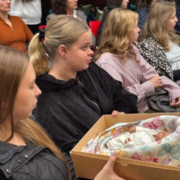 Renginio dalyvė sėdi auditorijoje, rankose laikydama dėžę su naujagimiui skirtomis priemonėmis.