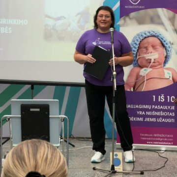 Violetinius marškinėlius vilkinti pranešėja kalba prie „Neišnešiotuko“ informacinio stendo.