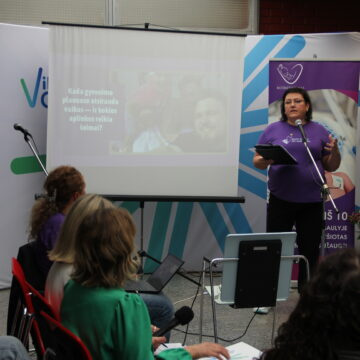 Pranešėja violetiniais marškinėliais pristato informaciją auditorijai prie projekcinio ekrano.