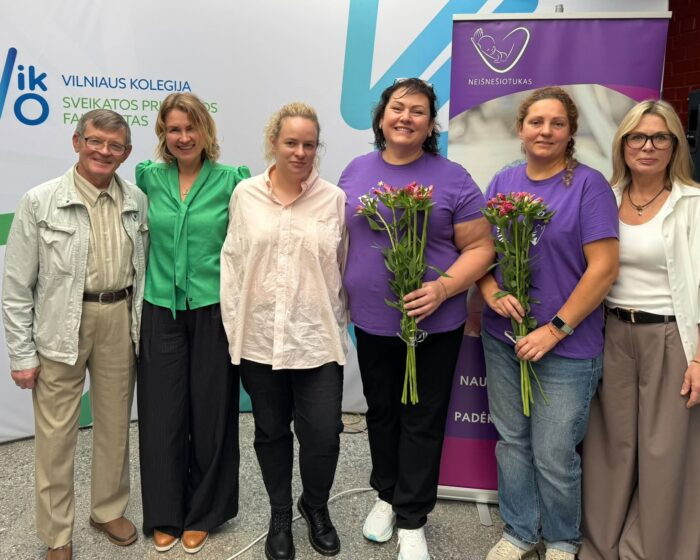 Šešios renginio dalyvės pozuoja bendrai nuotraukai prie organizacijų stendų.